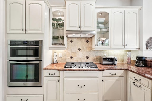 kitchen with under cabinet range hood, stainless steel appliances, glass insert cabinets, and backsplash