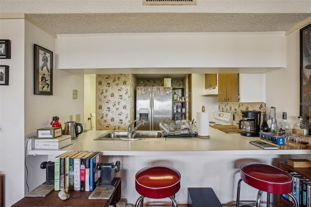 kitchen with under cabinet range hood, a sink, a textured ceiling, a peninsula, and stainless steel fridge with ice dispenser