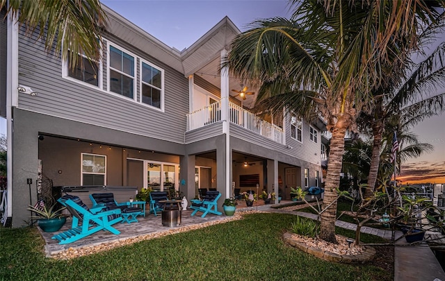 back of house at dusk with a balcony, stucco siding, a lawn, and a patio