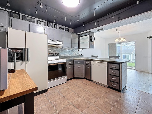 kitchen featuring white appliances, a peninsula, decorative backsplash, under cabinet range hood, and dark countertops