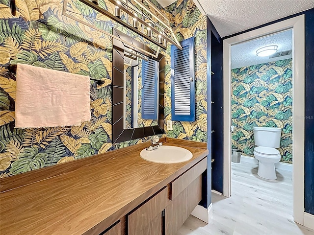 bathroom featuring vanity, wood finished floors, wallpapered walls, a textured ceiling, and toilet