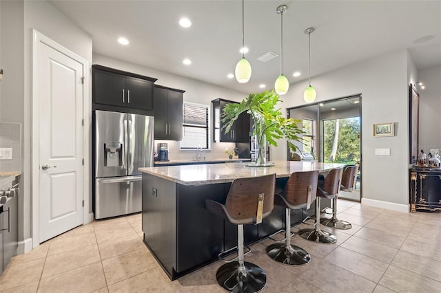 kitchen featuring a center island, light stone counters, stainless steel refrigerator with ice dispenser, light tile patterned flooring, and dark cabinets