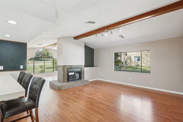 living room featuring visible vents, baseboards, vaulted ceiling with beams, a brick fireplace, and light wood-type flooring