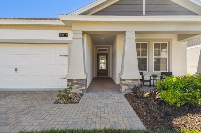 doorway to property featuring a porch, stucco siding, decorative driveway, a garage, and stone siding