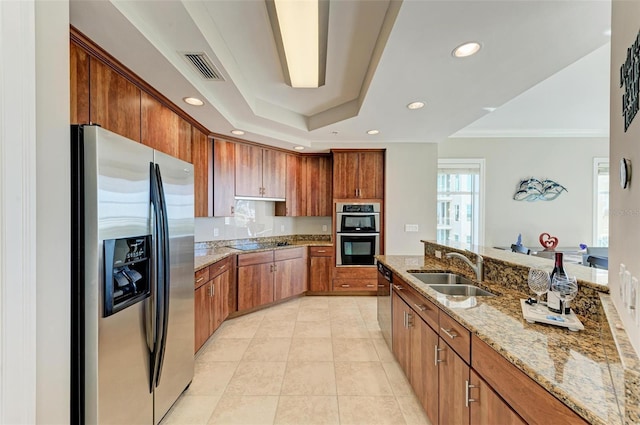 kitchen featuring light tile patterned floors, appliances with stainless steel finishes, brown cabinets, a tray ceiling, and a sink