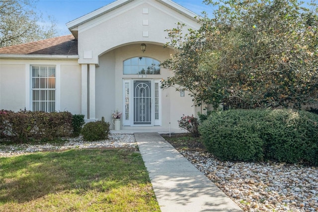 doorway to property with a yard, roof with shingles, and stucco siding