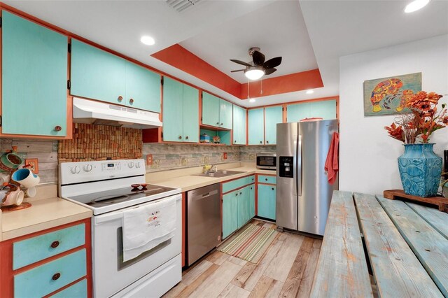 kitchen with light wood-style flooring, under cabinet range hood, stainless steel appliances, a sink, and a raised ceiling