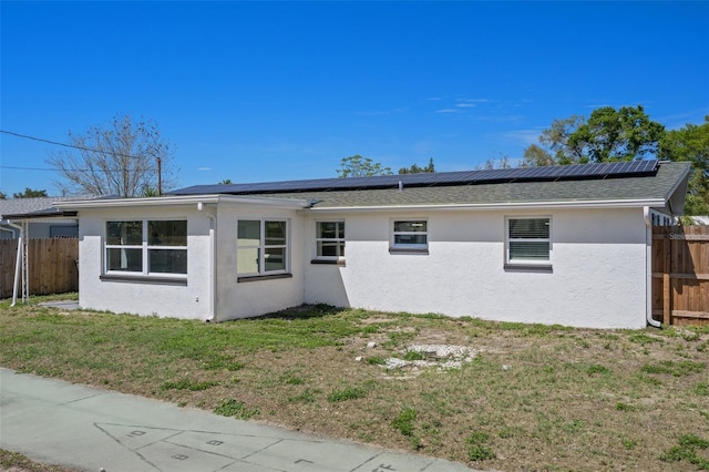 rear view of house featuring stucco siding, a lawn, roof mounted solar panels, fence, and roof with shingles