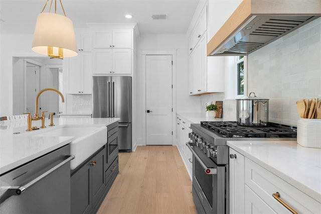 kitchen with premium appliances, white cabinets, hanging light fixtures, light wood-type flooring, and wall chimney exhaust hood