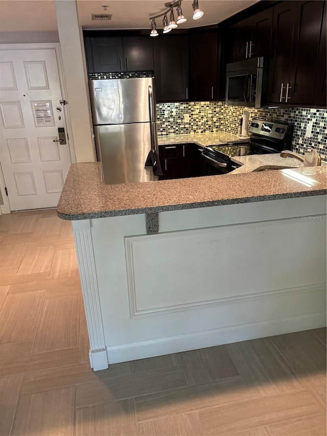 kitchen featuring light stone counters, stainless steel appliances, backsplash, dark cabinets, and a peninsula