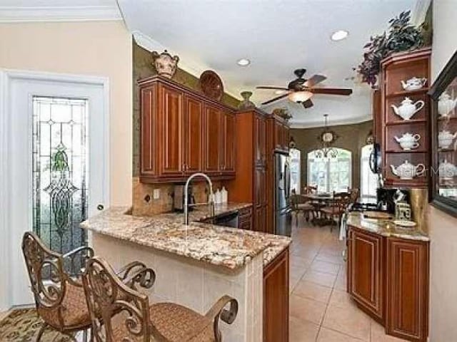 kitchen with brown cabinetry, ornamental molding, freestanding refrigerator, light stone countertops, and light tile patterned flooring