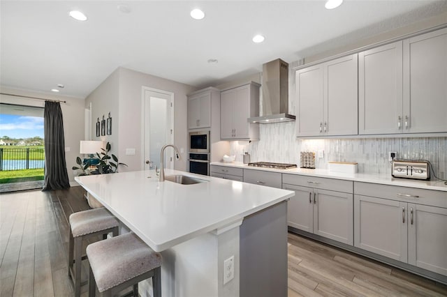 kitchen with a center island with sink, decorative backsplash, gray cabinetry, a sink, and wall chimney exhaust hood