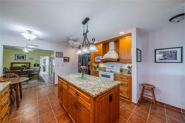 kitchen featuring decorative light fixtures, brown cabinetry, a kitchen island, white appliances, and premium range hood