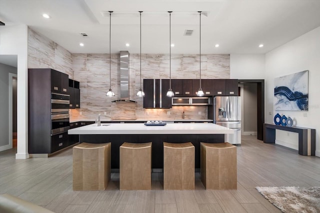 kitchen with visible vents, wall chimney range hood, light countertops, a large island with sink, and appliances with stainless steel finishes