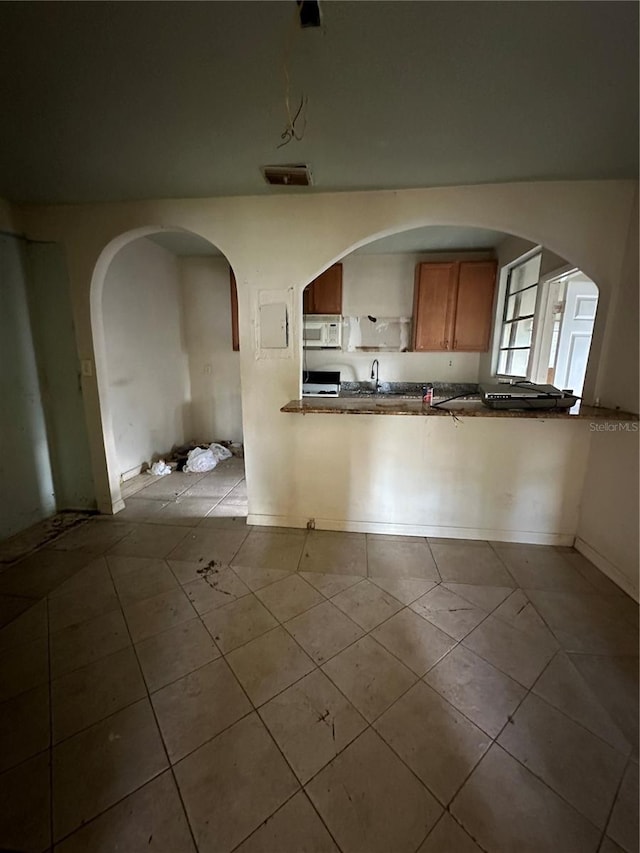 kitchen with brown cabinetry, dark countertops, white microwave, tile patterned floors, and a peninsula