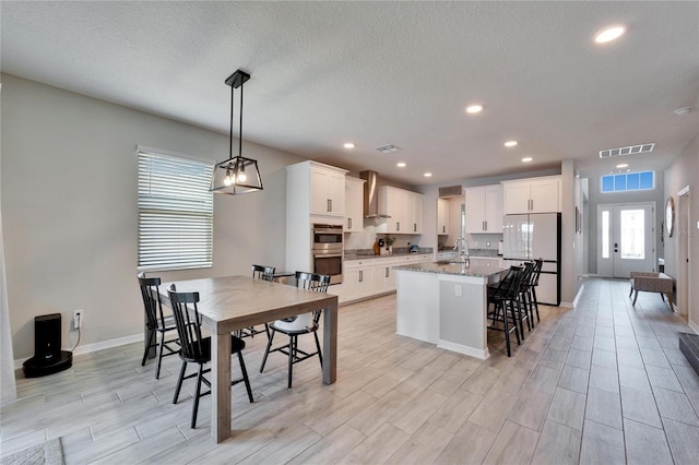 kitchen with visible vents, an island with sink, a sink, freestanding refrigerator, and wall chimney exhaust hood