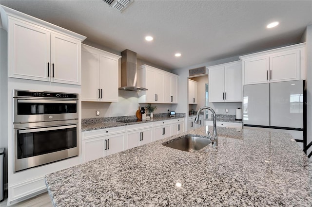 kitchen with visible vents, stainless steel double oven, a sink, white cabinetry, and wall chimney range hood