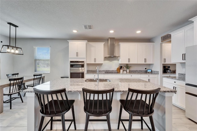 kitchen featuring visible vents, refrigerator, a kitchen island with sink, a sink, and wall chimney exhaust hood