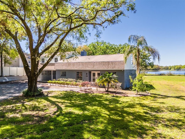 ranch-style house with a front lawn, fence, a water view, roof with shingles, and a chimney