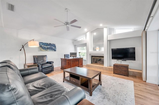 living area with a ceiling fan, visible vents, light wood-style flooring, vaulted ceiling, and a glass covered fireplace