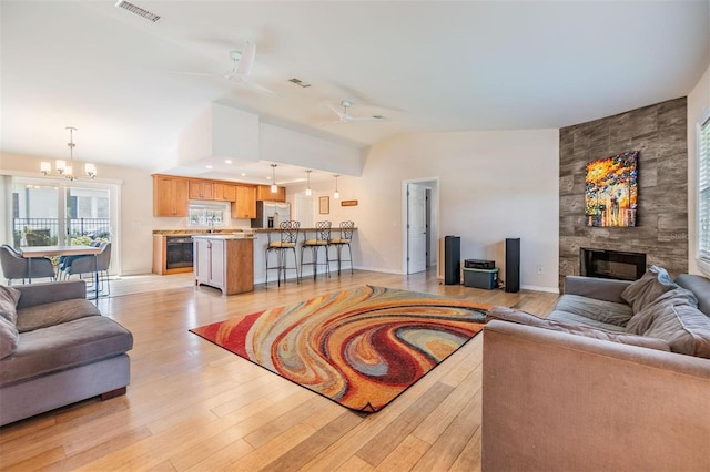 living room featuring lofted ceiling, a large fireplace, ceiling fan with notable chandelier, visible vents, and light wood finished floors