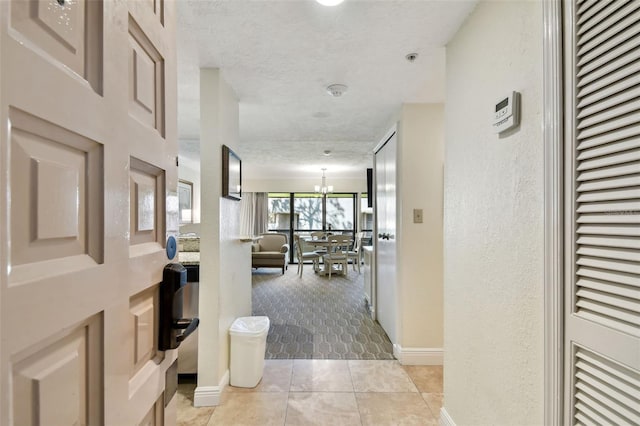 hallway featuring a chandelier, a textured wall, light tile patterned floors, a textured ceiling, and baseboards