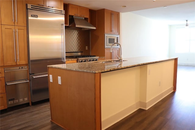 kitchen featuring dark wood-style floors, built in appliances, light stone countertops, under cabinet range hood, and a sink