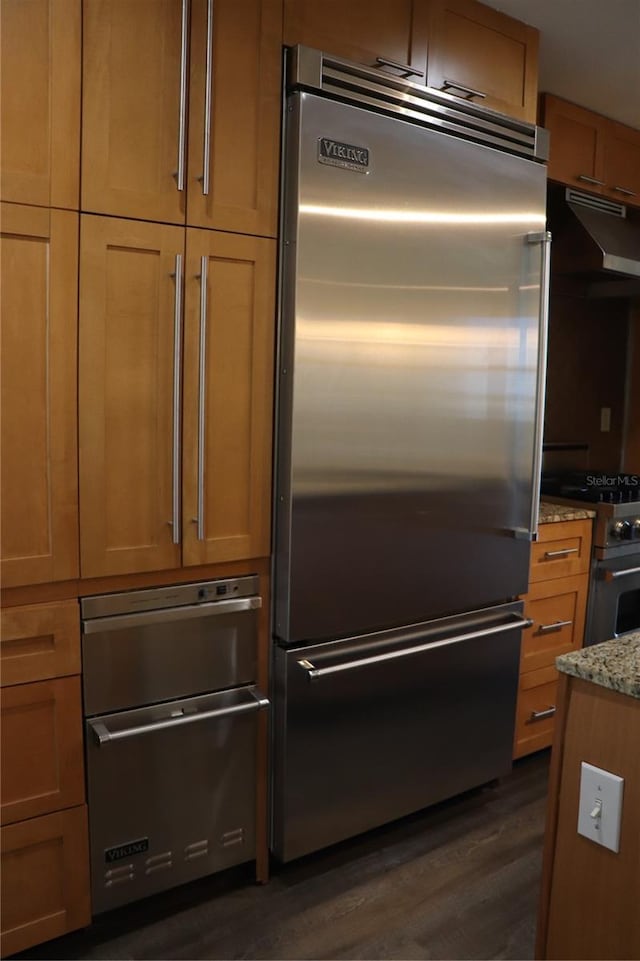 kitchen with high end appliances, dark wood finished floors, light stone countertops, and under cabinet range hood