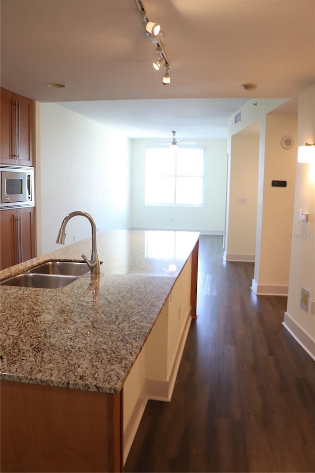 kitchen featuring brown cabinets, stainless steel microwave, dark wood-type flooring, a kitchen island with sink, and a sink