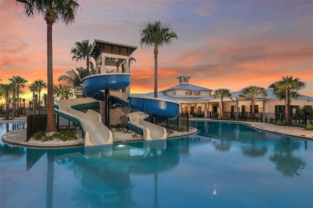 pool at dusk with playground community, a water slide, a community pool, and fence