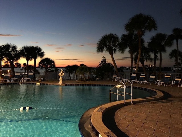 pool at dusk with a patio area, fence, and a community pool