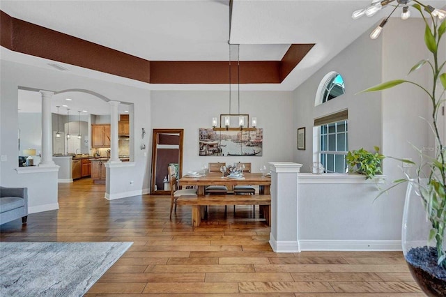 dining space with a raised ceiling, a notable chandelier, light wood-style flooring, decorative columns, and baseboards