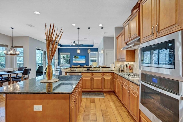 kitchen with brown cabinetry, dark stone counters, stainless steel appliances, ceiling fan with notable chandelier, and light wood-type flooring