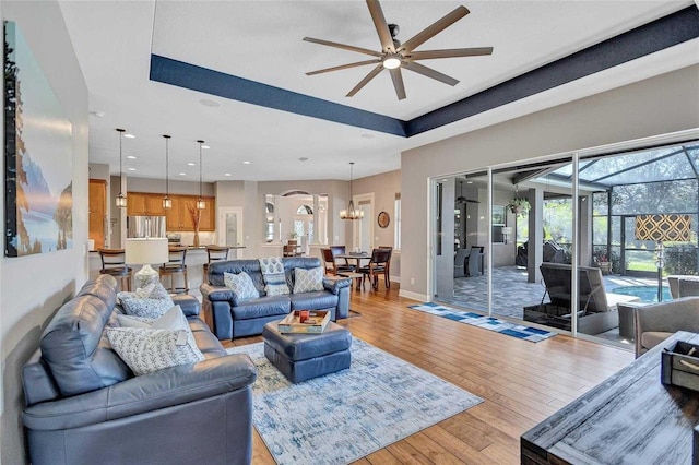 living room with light wood finished floors, baseboards, recessed lighting, ceiling fan with notable chandelier, and a raised ceiling