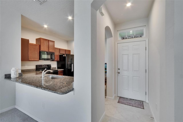 kitchen with dark stone countertops, arched walkways, brown cabinetry, black appliances, and a sink