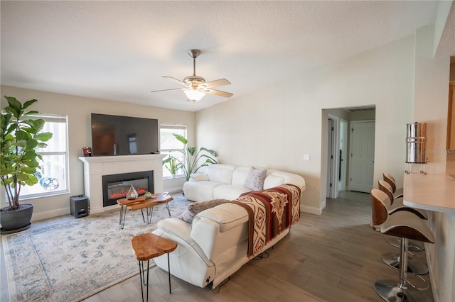 living area featuring a glass covered fireplace, baseboards, wood finished floors, and ceiling fan