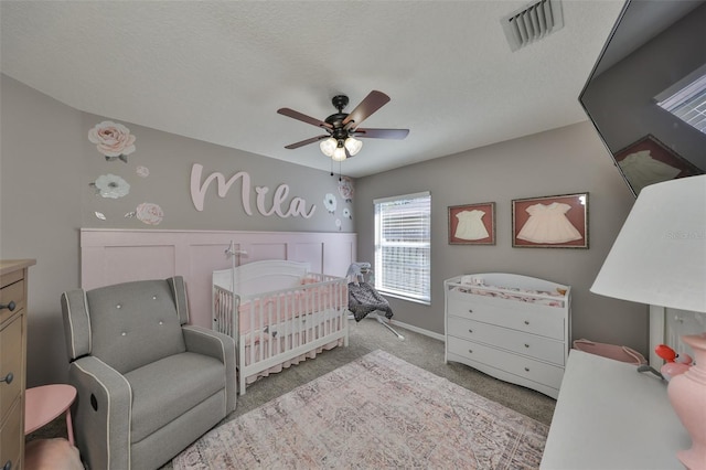 carpeted bedroom with visible vents, a nursery area, a ceiling fan, and a textured ceiling