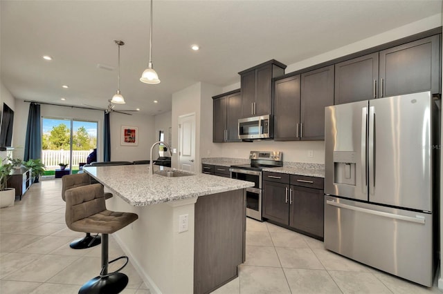 kitchen with an island with sink, a sink, light stone counters, appliances with stainless steel finishes, and light tile patterned floors