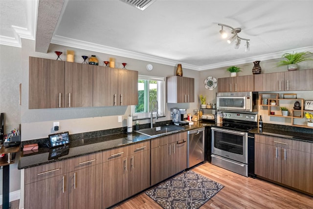 kitchen with light wood finished floors, ornamental molding, dark stone countertops, appliances with stainless steel finishes, and a sink