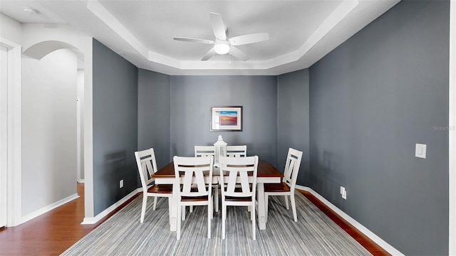 dining area with arched walkways, a raised ceiling, ceiling fan, wood finished floors, and baseboards