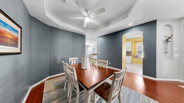 dining room featuring arched walkways, a textured ceiling, wood finished floors, baseboards, and a tray ceiling