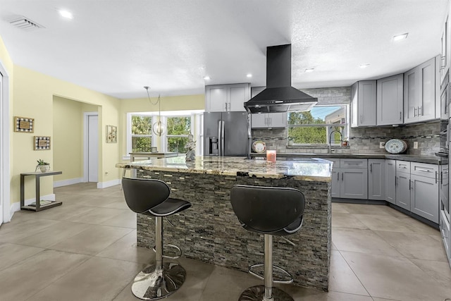 kitchen with plenty of natural light, island exhaust hood, gray cabinetry, stainless steel fridge, and tasteful backsplash