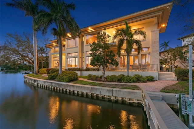 back of house at twilight with stucco siding, a balcony, and a water view