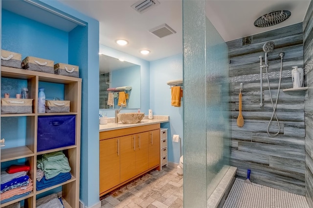 full bathroom featuring visible vents, a tile shower, brick floor, baseboards, and vanity