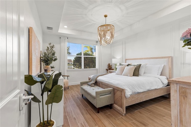 bedroom featuring wood finish floors, visible vents, a decorative wall, a raised ceiling, and a chandelier