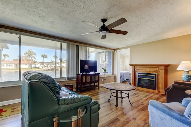 living room with light wood-type flooring, a textured ceiling, a glass covered fireplace, baseboards, and ceiling fan