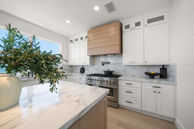 kitchen featuring premium range hood, visible vents, high end stove, white cabinets, and decorative backsplash