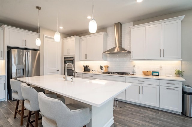 kitchen featuring a kitchen island with sink, stainless steel appliances, a kitchen bar, wall chimney range hood, and backsplash