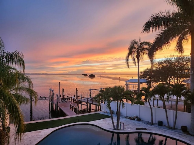 pool at dusk with a water view, boat lift, a dock, and fence
