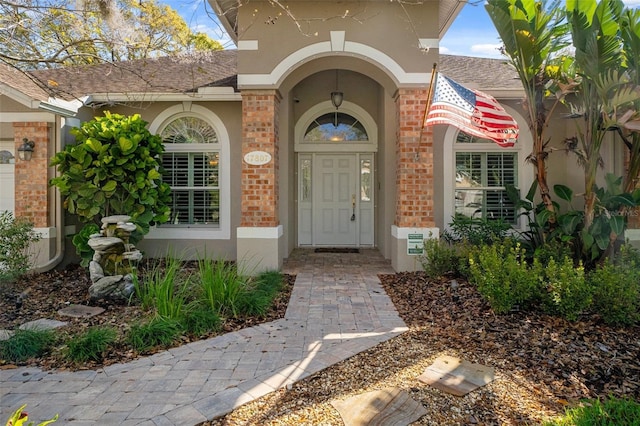 view of exterior entry featuring brick siding, roof with shingles, and stucco siding